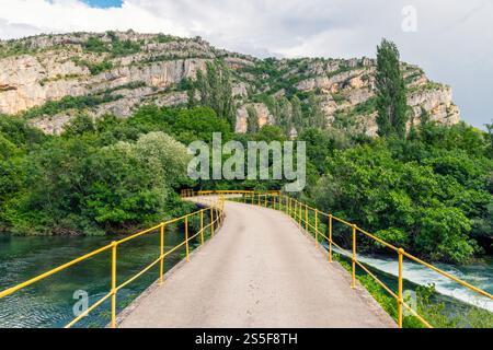 Yellow railed bridge over a river with lush greenery and towering cliffs in the background under a cloudy sky, Krka national park Stock Photo