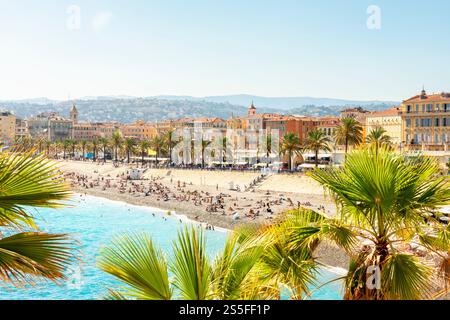 Sunbathers enjoying a sunny day on busy beach of Nice with clear blue waters, palm trees in the foreground, and historic coastal buildings in the background, Nice, France Stock Photo