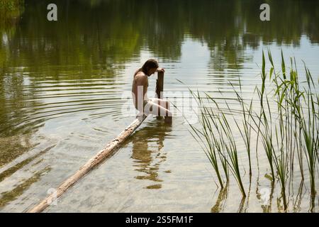 Barefoot man in shorts sitting on a log over a tranquil lake surrounded by greenery. Stock Photo