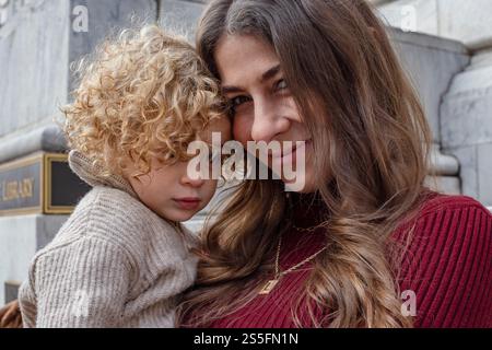 A curly-haired child nestled in the embrace of a smiling woman with long hair against a stone wall backdrop. Cleveland, Ohio, USA Stock Photo