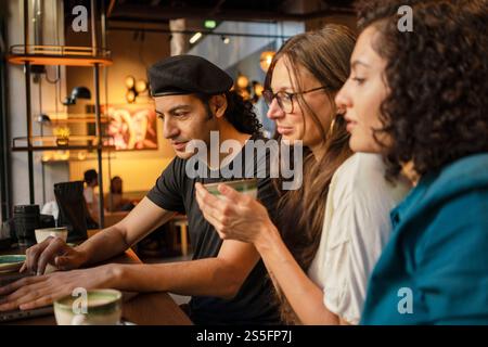 Three adults sit at a cafe table, one man working on a laptop and two women engaged in a conversation, with coffee cups in front of them, Berlin, Germany Stock Photo