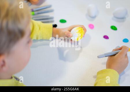Caucasian child painting easter eggs with yellow paint on a white table, using colorful markers Stock Photo