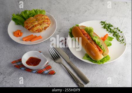 Appetizing sausage bread roll in white dish and large hotdog on wooden cutting board, tomato sauce, fork and knife on floor, copy space Stock Photo