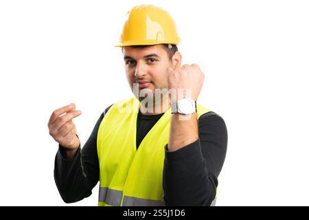 Adult builder man in work protective equipment helmet and vest showing time means money gesture with fingers and presenting watch isolated on white ba Stock Photo