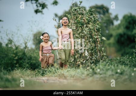 Portrait of two Cute girls in Thai traditional dress and put white flower on her ear walking hand in hand on rice field, They are smile with happiness Stock Photo