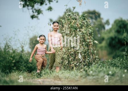 Portrait of two Cute girls in Thai traditional dress and put white flower on her ear walking hand in hand on rice field, They are smile with happiness Stock Photo