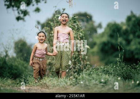 Portrait of two Cute girls in Thai traditional dress and put white flower on her ear walking hand in hand on rice field, They are smile with happiness Stock Photo