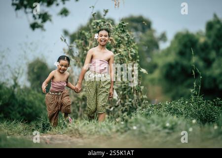 Portrait of two Cute girls in Thai traditional dress and put white flower on her ear walking hand in hand on rice field, They are smile with happiness Stock Photo