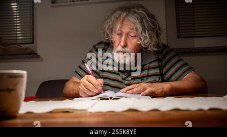An older man with curly hair writes in a notebook under warm indoor lighting, deeply engrossed in his thoughts, with a cup nearby. Stock Photo