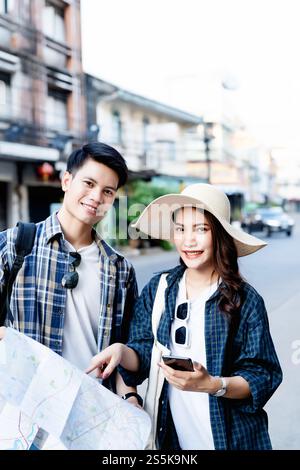 Young Backpacker Couple Hold paper Map and smartphone to checking location direction On City Street, pretty woman wear sombrero, They looking at Stock Photo