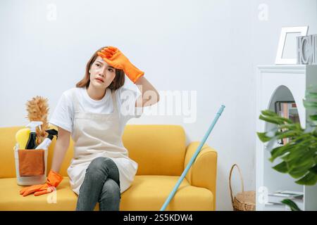 Young housekeeper woman sitting on sofa in living room with feel tired, She wipe sweat and take a break after finishing cleaning the house, copy space Stock Photo
