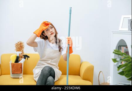 Young housekeeper woman sitting on sofa in living room with feel tired, She wipe sweat and take a break after finishing cleaning the house, copy space Stock Photo