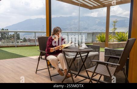 Senior woman with glasses reading a book on a garden terrace during the morning with mountains in the background Stock Photo