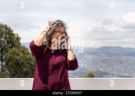 Senior woman with glasses holds her windblown hair while talking on cell phone outdoors Stock Photo
