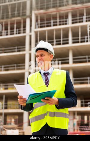 Foreman in protective helmet and vest with folder of documents on construction site Stock Photo