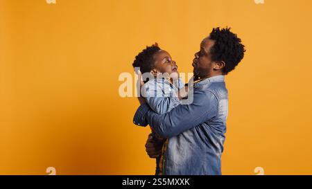 African american dad caressing and kissing his adorable baby boy in studio, carrying his toddler and playing around with him. Cute family showing love and devotion to his infant son. Camera B. Stock Photo