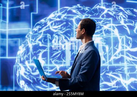Computer scientist analyzes AI models with large digital brain displayed behind. IT specialist in workspace using machine learning to better understand and develop artificial intelligence Stock Photo