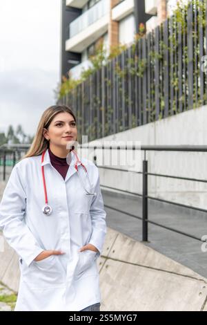 Young blonde Latina female doctor walks with her apron and stethoscope down the street next to a building Stock Photo