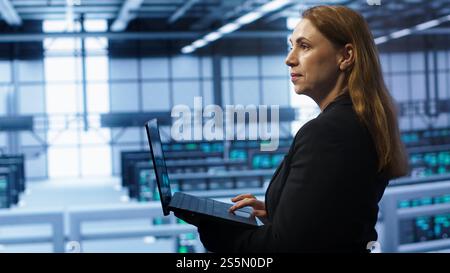 Computer scientist doing yearly maintenance using laptop to prolong data center equipment life span. IT specialist in server hub facility monitoring server infrastructure using device, camera A Stock Photo