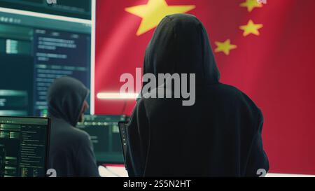 Chinese hacker working in governmental hacking room with Chinese flag on a big screen. Having the mission on inflicting fear and misinformation among the population. Dictatorship propaganda. Camera A. Stock Photo