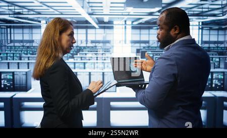 Teamworking coworkers doing maintenance using devices, prolonging data center hardware life span. Colleagues doing brainstorming in server hub, monitoring server infrastructure using devices, camera B Stock Photo
