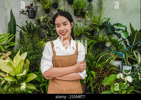 Portrait Young Asian gardener female wearing apron stand and cross arm, she smile and looking to camera, potted plant in background Stock Photo