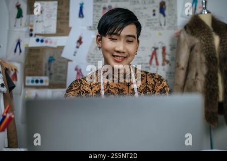 Close up face of Professional Asian young male tailor with measuring tape on neck working on laptop with drawings background in studio. Designer and Stock Photo