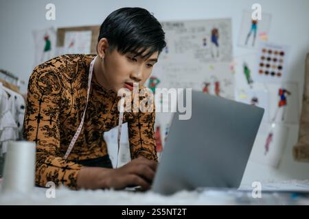 Portrait of Professional Asian young male tailor with measuring tape on neck working on laptop with drawings background in studio. Designer and LGBTQ Stock Photo