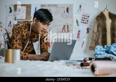 Portrait of Professional Asian young male tailor with measuring tape on neck working on laptop with drawings background in studio. Designer and LGBTQ Stock Photo