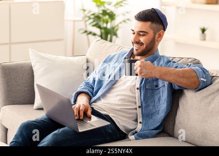 Smiling jewish man watching video on computer, drinking hot coffee Stock Photo