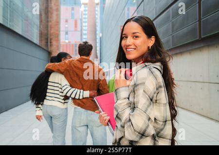 Cheerful student girl holding notebooks and headphones, smiling while her friends embrace each other, capturing the essence of youthful and academic life in a university campus or high school. High quality photo Stock Photo