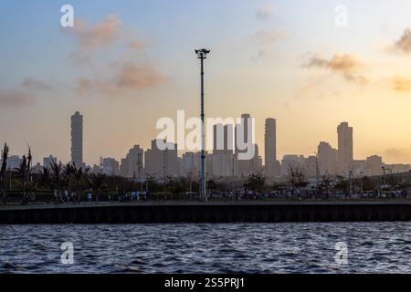 The Barranquilla skyline at sunset features high-rise buildings silhouetted against a golden sky, with a lively waterfront promenade and rippling wate Stock Photo