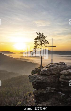 Sunrise on a rock with a summit cross and a view over a forest. Morning atmosphere with fog in the middle of nature. Landscape in the Palatinate Fores Stock Photo