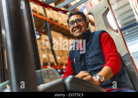 Happy latin worker smiling at camera while using forklift in a distribution warehouse Stock Photo