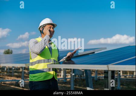 Asian Young engineer in white helmet holding checking board in hand standing and talking on smartphone while working at solar farm in background Stock Photo