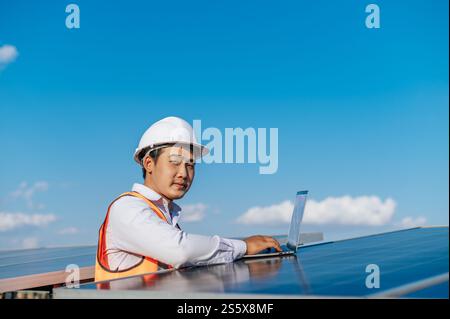 Young Asian Inspector Engineer man use laptop computer working at solar farm, Technician, supervisor male in white helmet Checking operation of sun Stock Photo