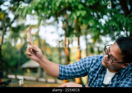 Portrait of Asian traveler man glasses pork steak frying, BBQ in roasting skillet pan or pot at a campsite. Outdoor cooking, traveling, camping, lifes Stock Photo