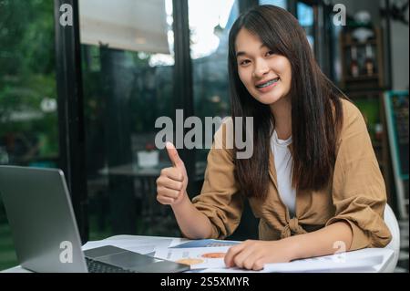 Young woman thumb up and smile while working on laptop and paperwork or report presenting Project growth graph at cafe Stock Photo
