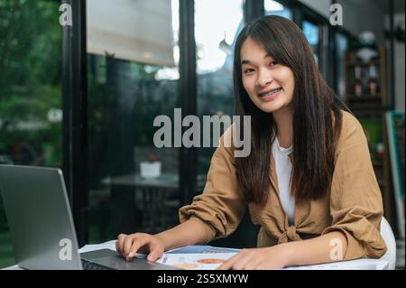 Portrait Young woman smile and looking to camera while working on laptop and paperwork or report presenting Project growth graph at cafe Stock Photo
