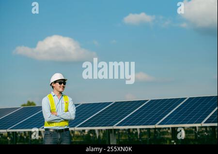 Portrait Asian Young engineer man wearing protective clothes, sunglasses and white helmet standing near solar panels while working in solar farm, Stock Photo