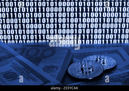 Two bitcoins lies on a pile of dollar bills on the background of a monitor depicting a binary code of bright zeros and one units on a black Stock Photo
