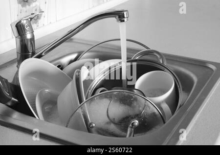 Dirty dishes and unwashed kitchen appliances lie in foam water under a tap from a kitchen faucet Stock Photo