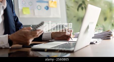 Elderly businessman taking notes while analyzing data on a laptop in a modern office Stock Photo
