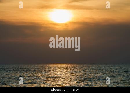 A tranquil sunset over the ocean, with the sun partially hidden by clouds. Warm golden light reflects on the calm sea, suggesting a tropical setting. Stock Photo