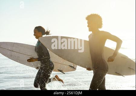 Sunset, beach and couple running in the water with surfboards to practice surfing while on vacation. Nature, sports and young man and woman surfers in Stock Photo