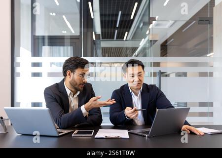 Two business colleagues discussing financial project while sitting at desk inside office, diverse people. Men serious and concentrated with laptops. Stock Photo