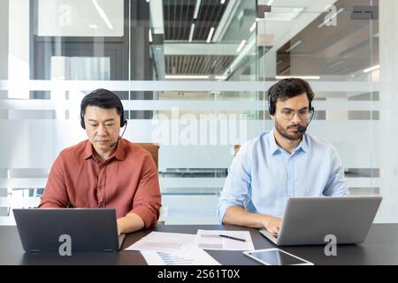 Two colleagues in casual clothes working inside office with laptops and headset. Office workers consulting clients remotely while sitting at desk. Stock Photo