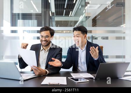 Two business colleagues behind paperwork rejoicing and celebrating victory triumph and successful achievement results. Men in business suits sitting at desk reviewing financial investment reports. Stock Photo