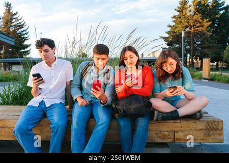 Young students using smartphones sitting on bench in university campus park Stock Photo