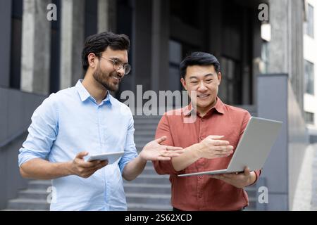 Two colleagues in casual clothes outside office building discussing current project on laptop. Office workers are focused and thinking, smiling contentedly Stock Photo
